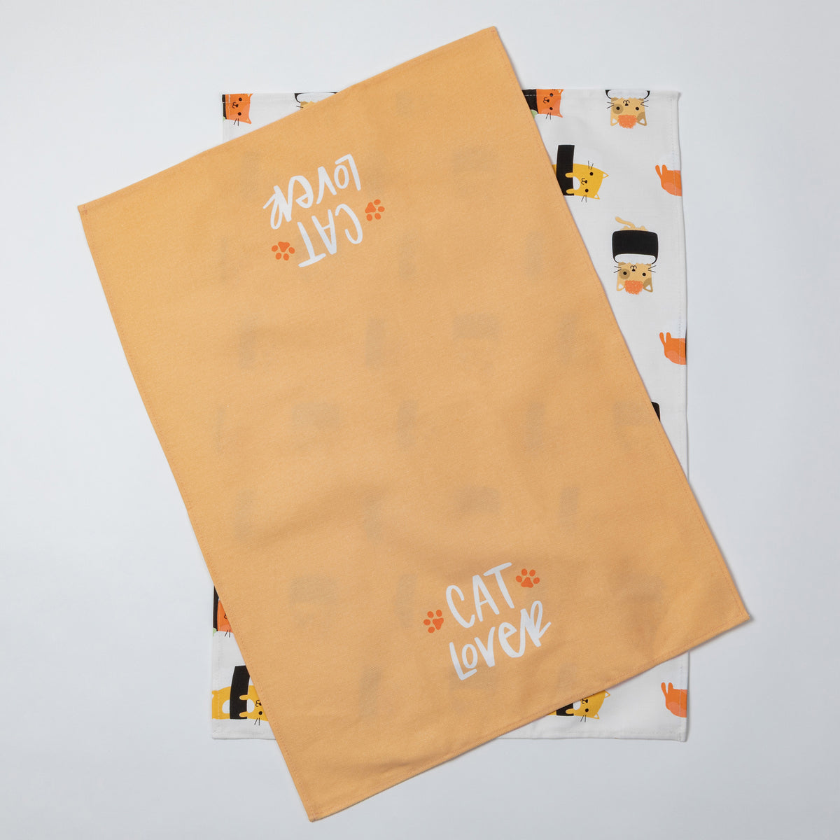 Two fabric tea towels laid flat on a light gray background. The top towel is a warm peachy-orange color with 'CAT LOVER' and small paw prints in white.