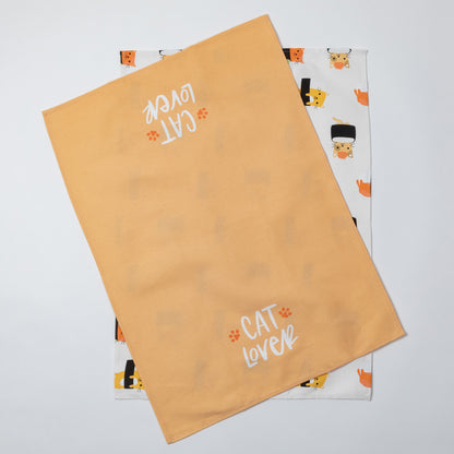 Two fabric tea towels laid flat on a light gray background. The top towel is a warm peachy-orange color with 'CAT LOVER' and small paw prints in white.