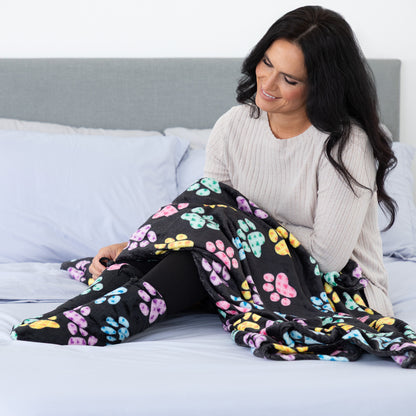 A woman sitting on a bed wearing a light beige ribbed sweater and black leggings, covered with a black fleece blanket featuring a colorful pattern of pink, yellow, blue, and purple paw prints.