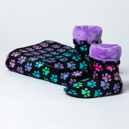 A matching pet fleece blanket and booties set on a light gray studio background. The set includes a black blanket and slippers covered in colorful paw print patterns in pink, purple, blue, and green, with plush purple trim on the slippers.