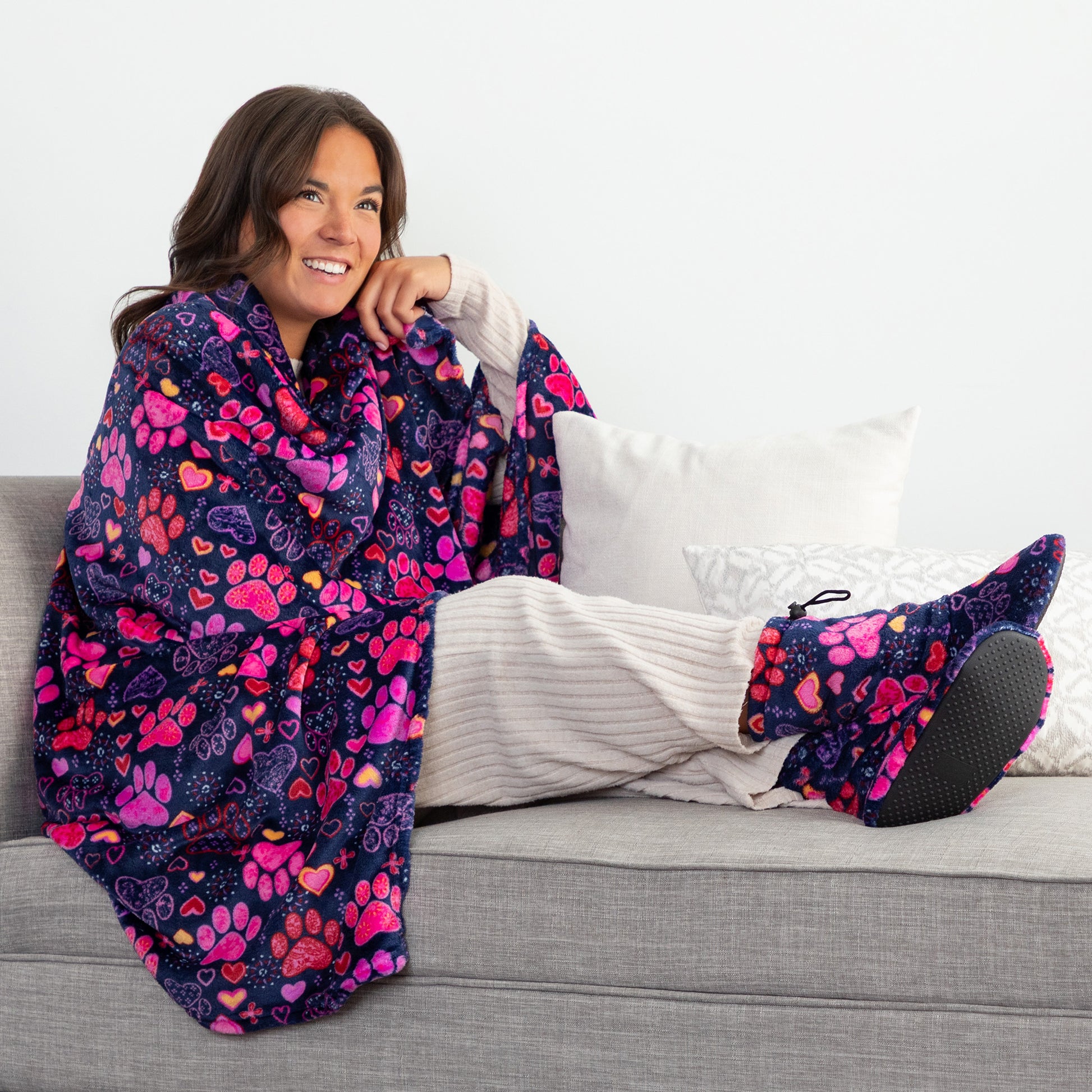 Woman sitting on a couch with a colorful paw print and heart blanket and matching fleece slippers.