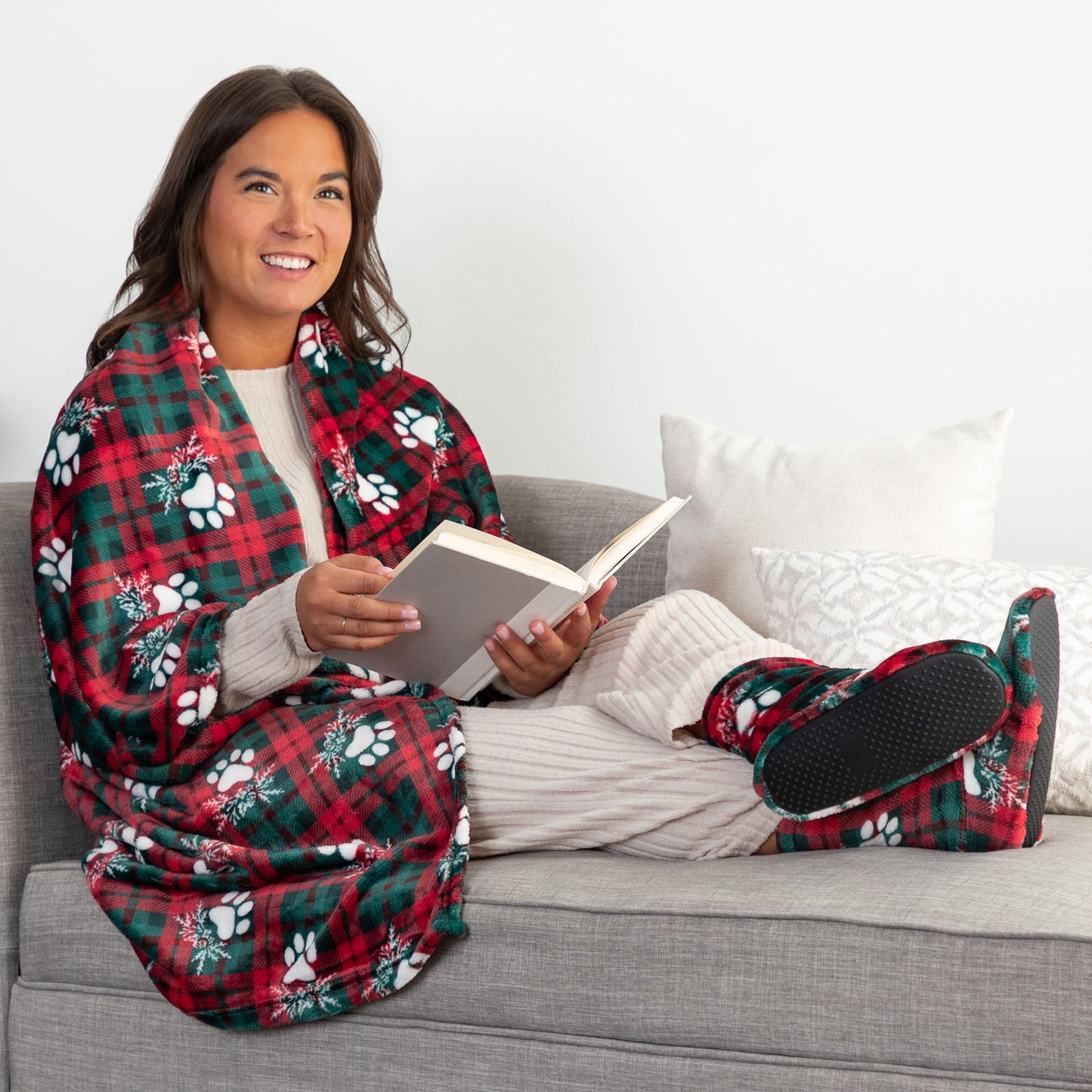 Woman sitting on a couch wearing a red and green plaid blanket with paw prints, holding a book.