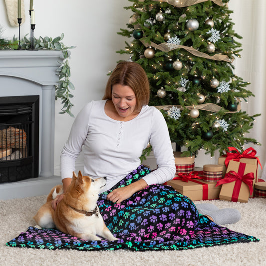 Woman sitting on the floor with a dog in front of a Christmas tree and presents with a rainbow paw blanket on lap.