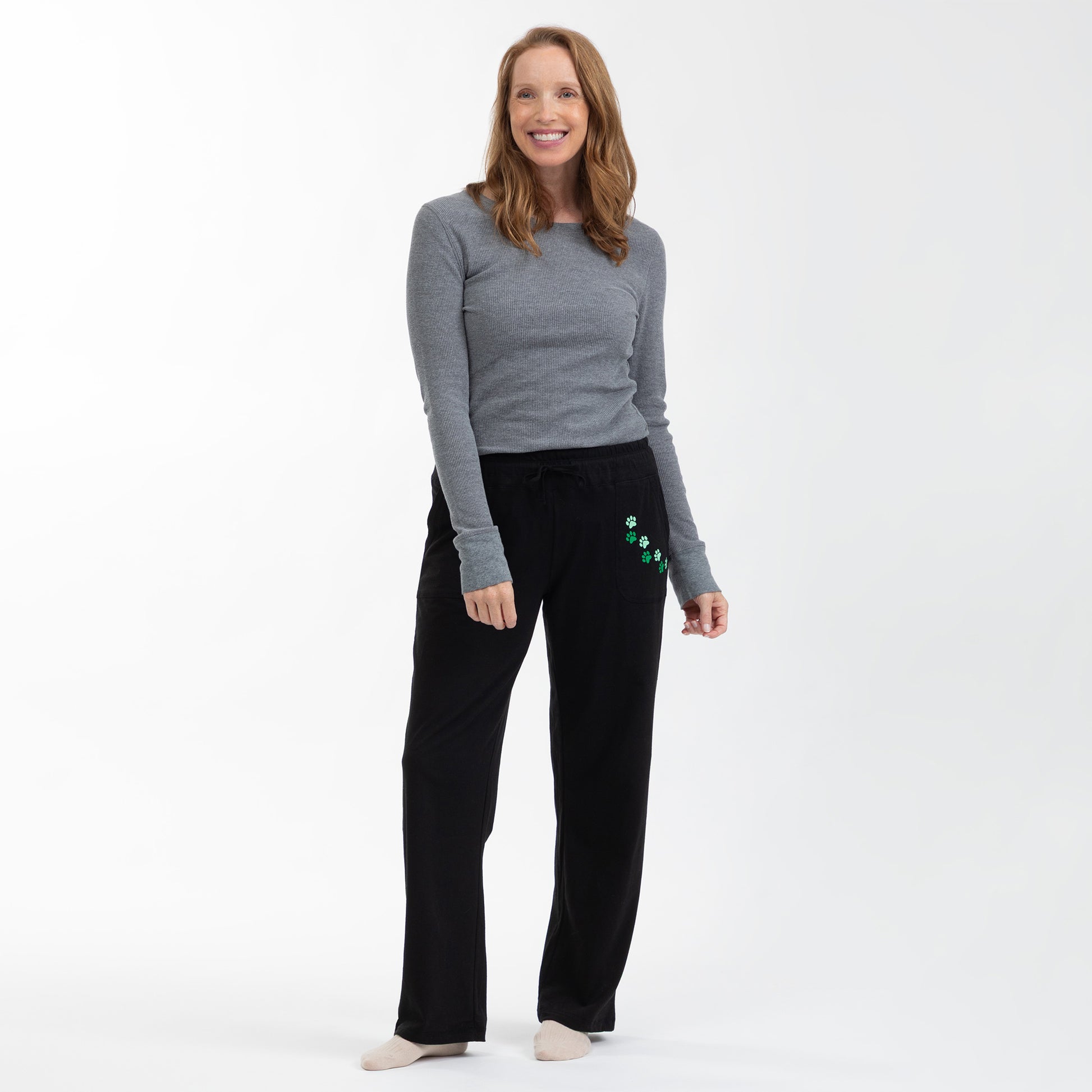A woman modeling black sweatpants with green paw print details on the pocket, paired with a gray long-sleeve top, against a plain white studio background.