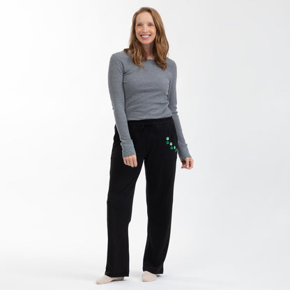 A woman modeling black sweatpants with green paw print details on the pocket, paired with a gray long-sleeve top, against a plain white studio background.