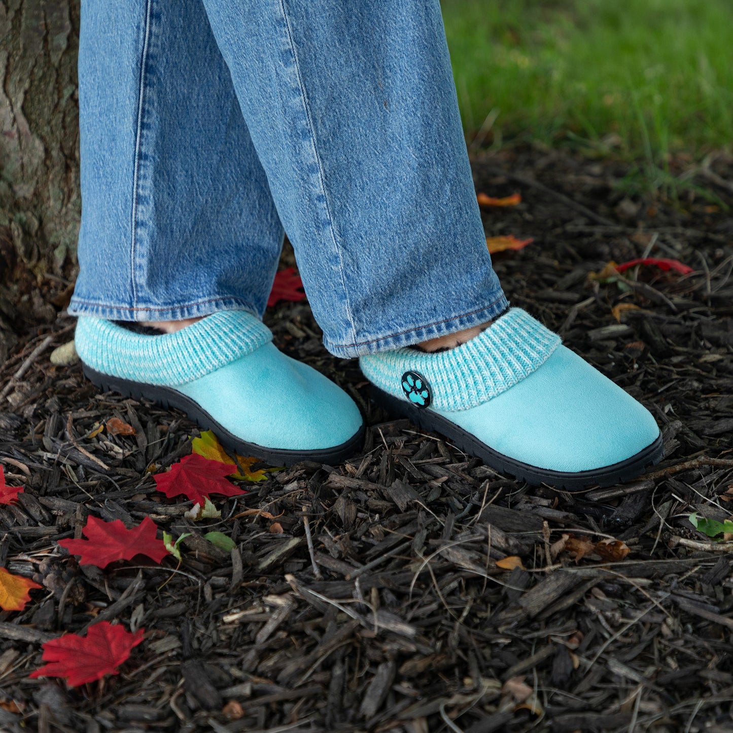 A person wearing light aqua slip-on slippers with ribbed knit cuffs and a small black paw print detail, standing outdoors on mulch near fallen leaves and tree bark.
