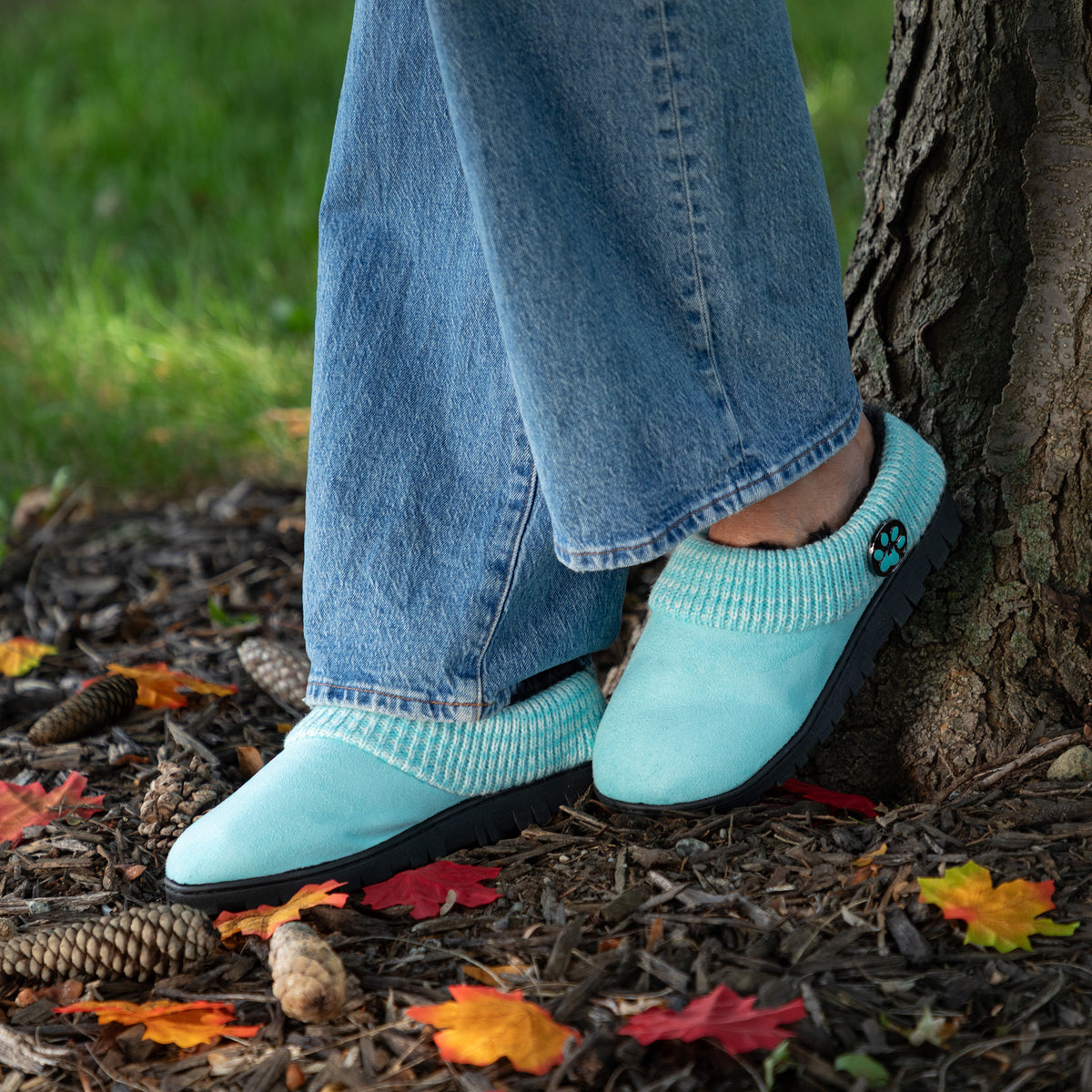 A person wearing light blue knit slip-on slippers with black soles, standing outdoors against a tree trunk on mulch with fallen leaves.