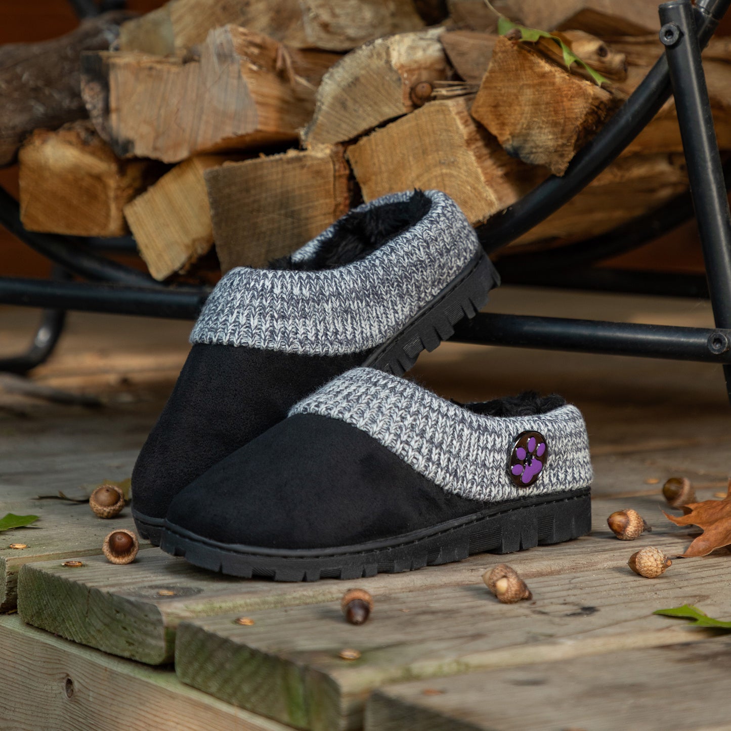 Black and gray slippers with a paw print logo on a wooden surface with firewood in the background.