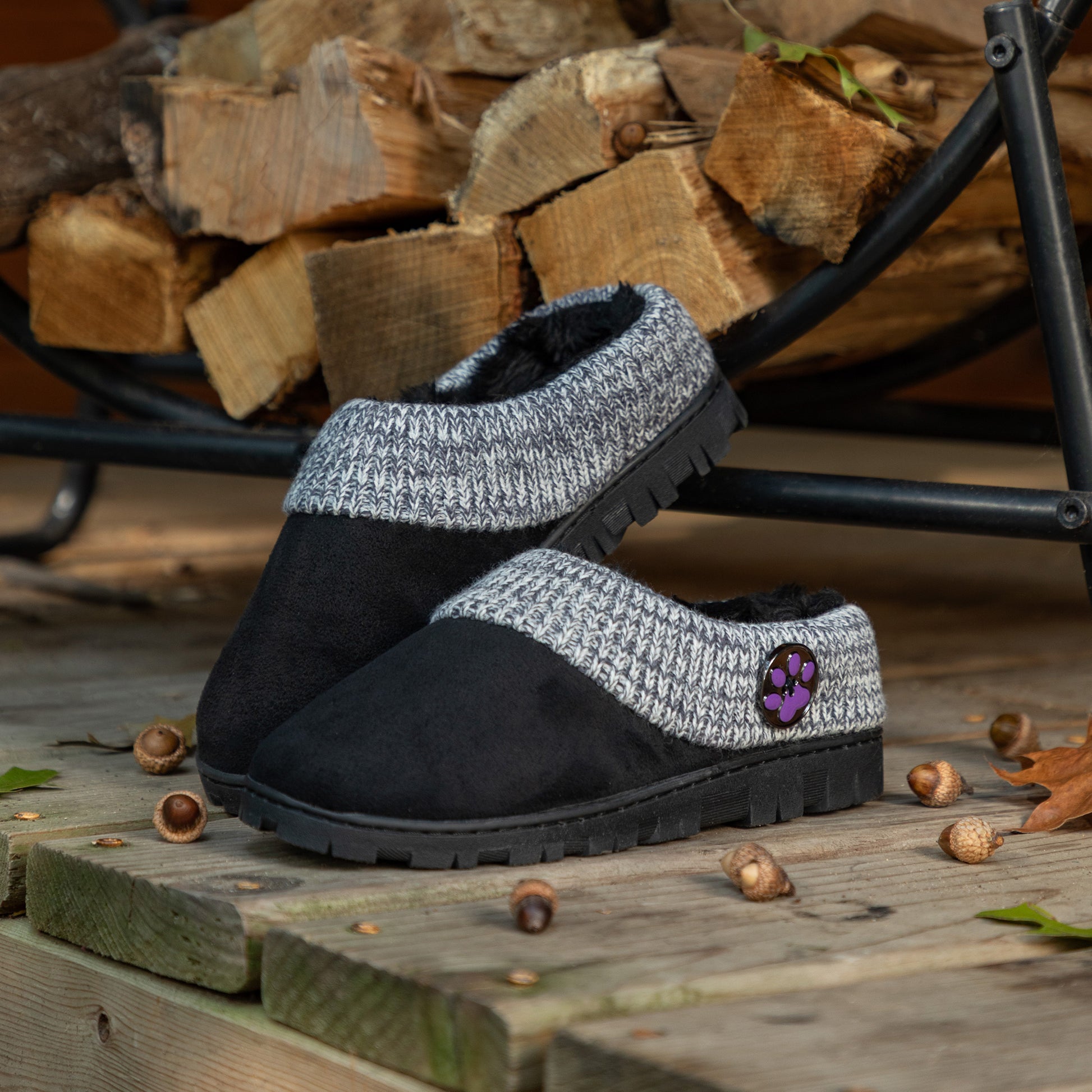 Black and gray slippers with a paw print logo on a wooden surface with firewood in the background.