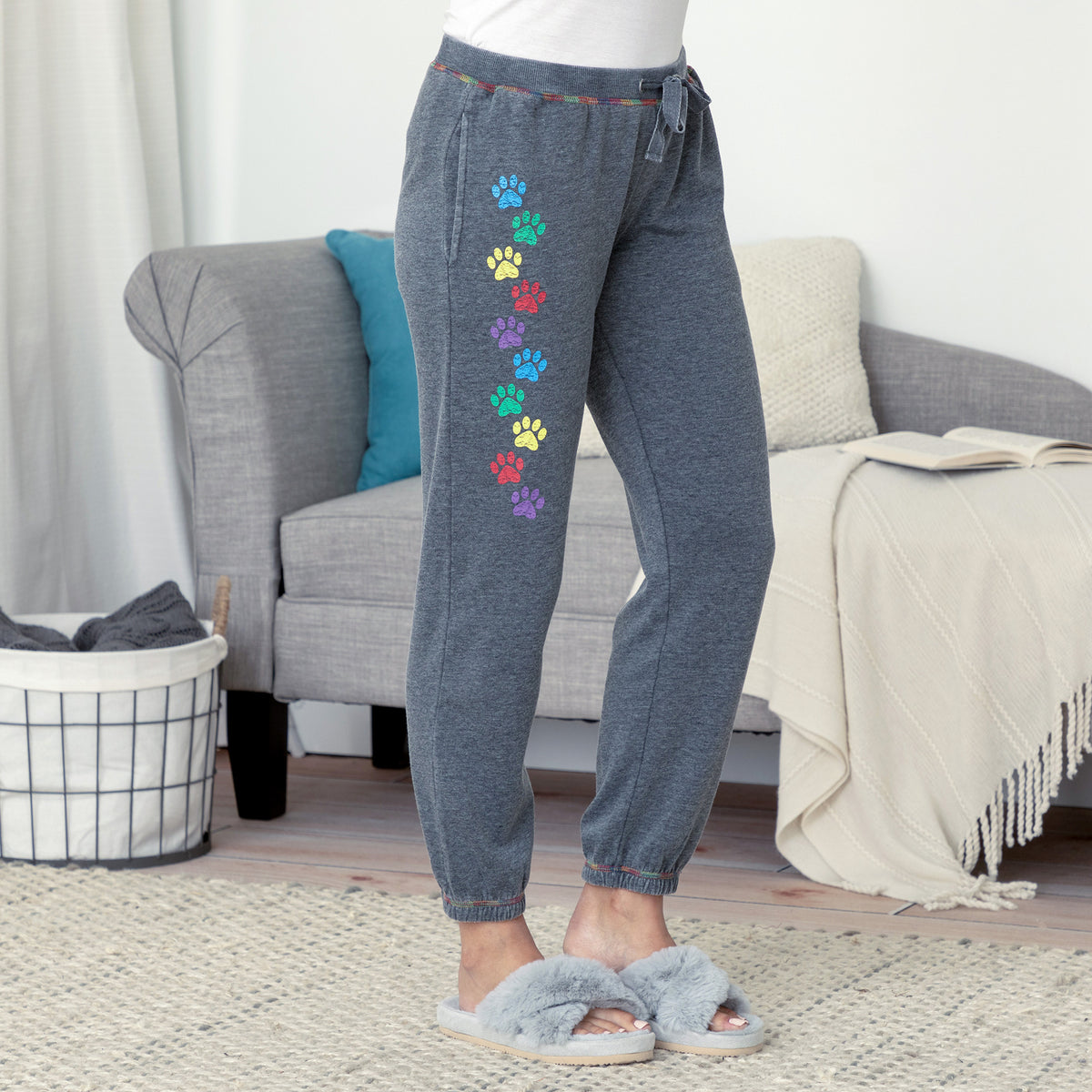 A person wearing dark heather gray jogger-style pants with a vertical row of colorful paw prints down one leg, standing in a living room with a gray sofa, throw blanket, and slippers.