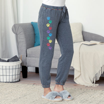 A person wearing dark heather gray jogger-style pants with a vertical row of colorful paw prints down one leg, standing in a living room with a gray sofa, throw blanket, and slippers.