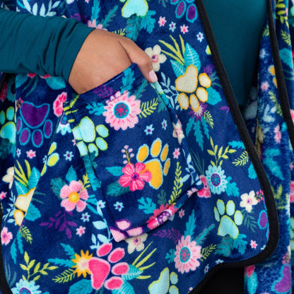 Hands in the pockets of a blue fleece shawl, patterned with paws, flowers and foliage. The edges are lined with black piping.