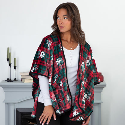 Woman in a cozy setting wearing a fleece, pocketed shawl, patterned with red and green plaid overlayed with white paws and mistletoe and black piping, wrapped around her shoulders.