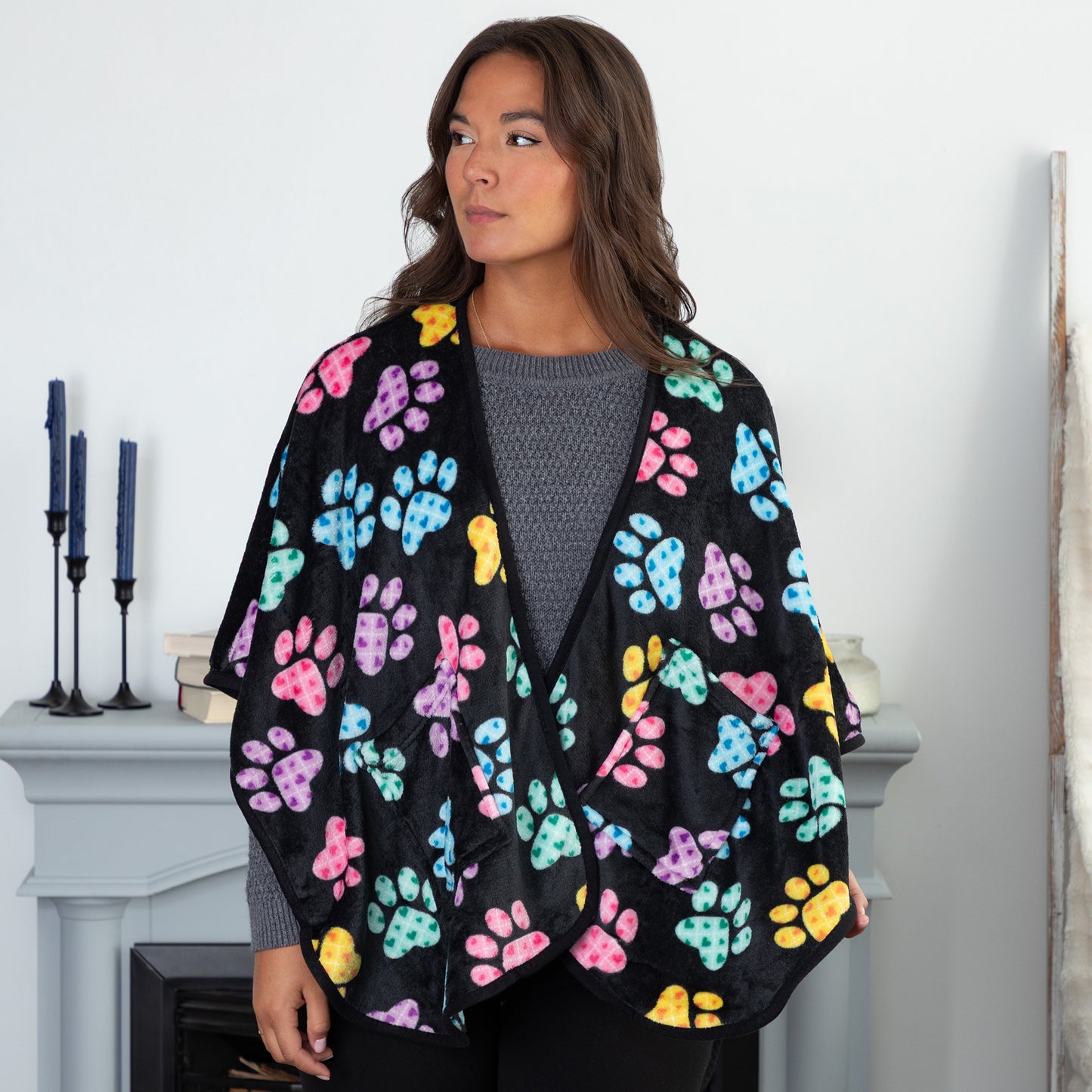 A woman standing in a cozy living room. She is wearing a black fleece, pocketed shawl, patterned with pink, blue, yellow, green, and purple argyle paw prints.