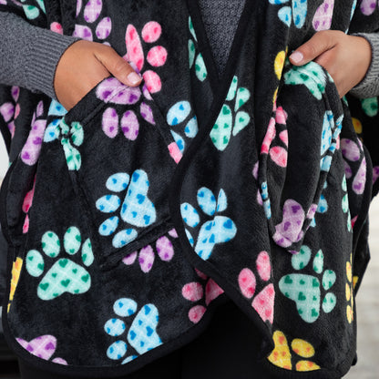 Hands in the two pockets of a black fleece wearable blanket shawl patterned with pink, blue, yellow, green, and purple argyle paw prints.