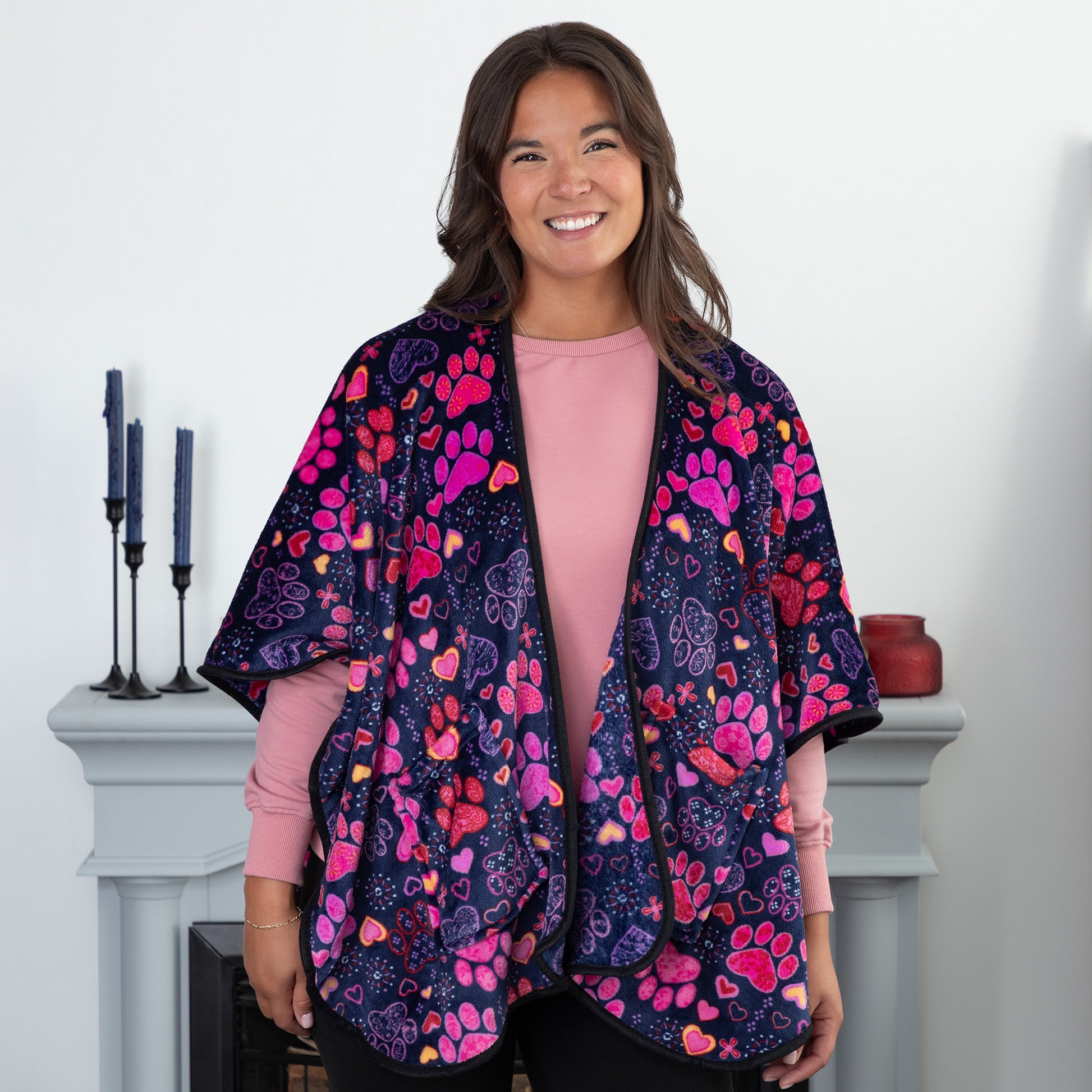 A woman standing in a cozy living room. She is wearing a deep blue based fleece, pocketed shawl, patterned with pink, red and purple paw prints paired with yellow, red, pink, and purple hearts. 
