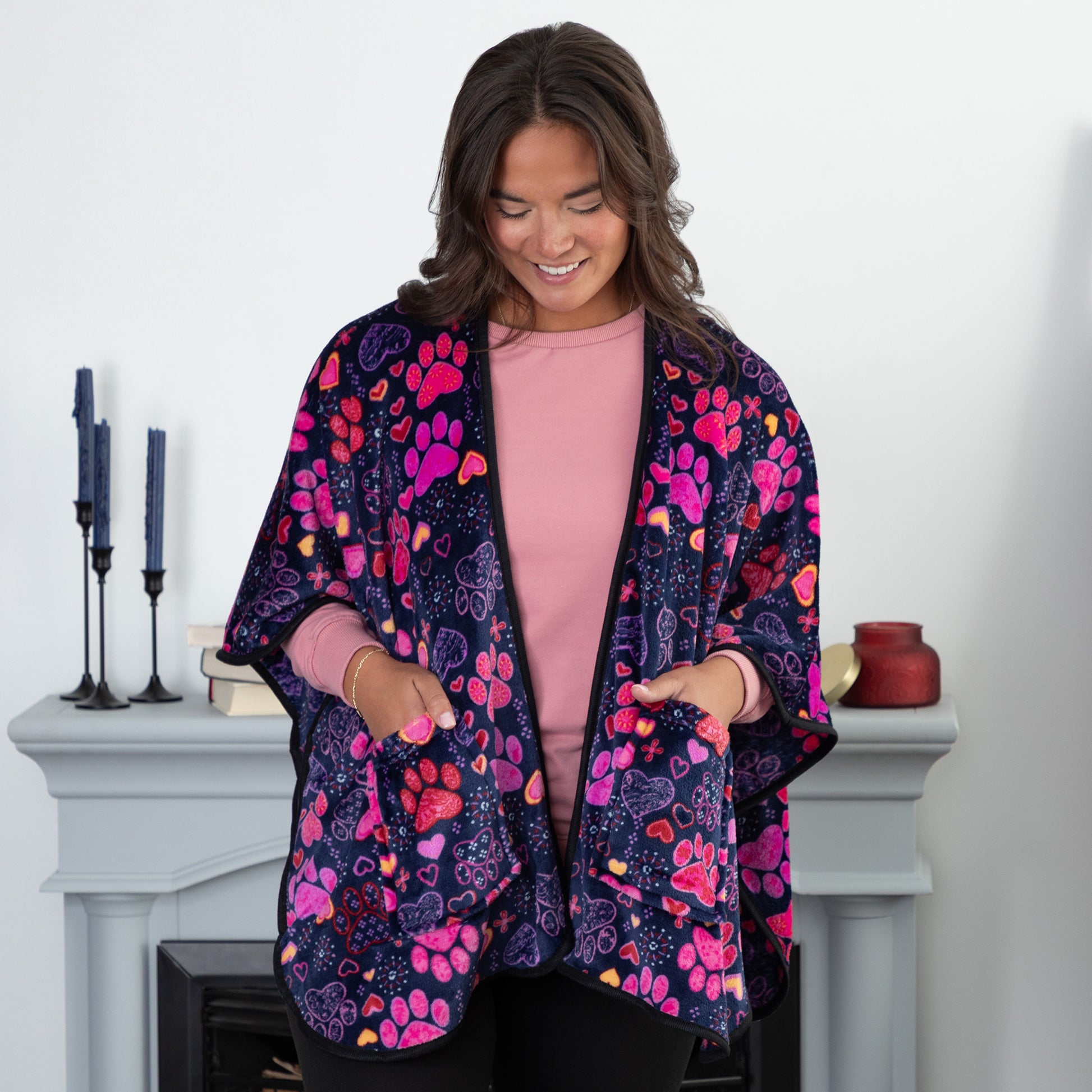 A woman with her hands in her pockets standing in a cozy living room. She is wearing a deep blue based fleece, pocketed shawl, patterned with pink, red and purple paw prints paired with yellow, red, pink, and purple hearts. 
