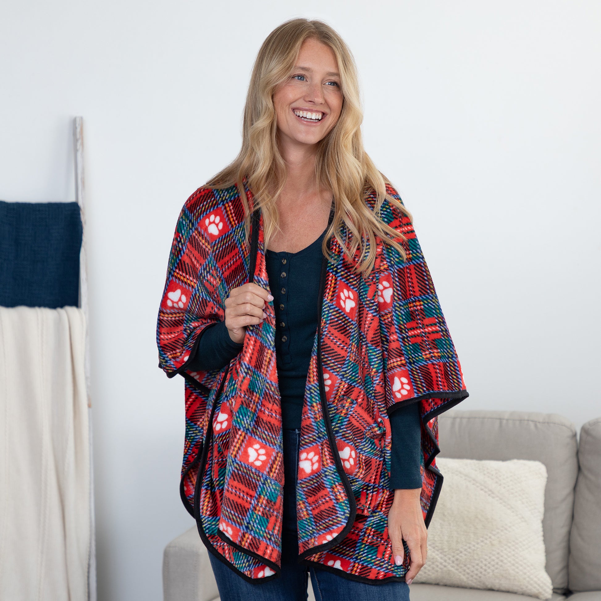 A woman stands indoors wearing a red, navy, and teal plaid poncho-style wrap with white paw print motifs over a black top. A sofa and neutral wall are visible in the background.