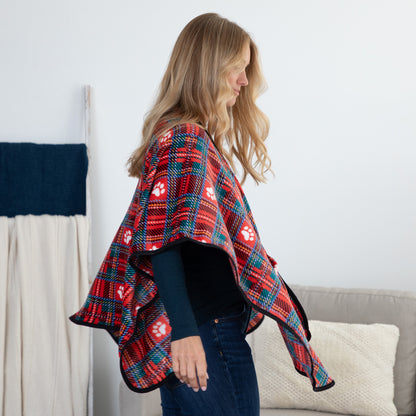 A woman wearing a red, teal, and white plaid blanket-style cape with paw print patterns, standing in a living room setting.