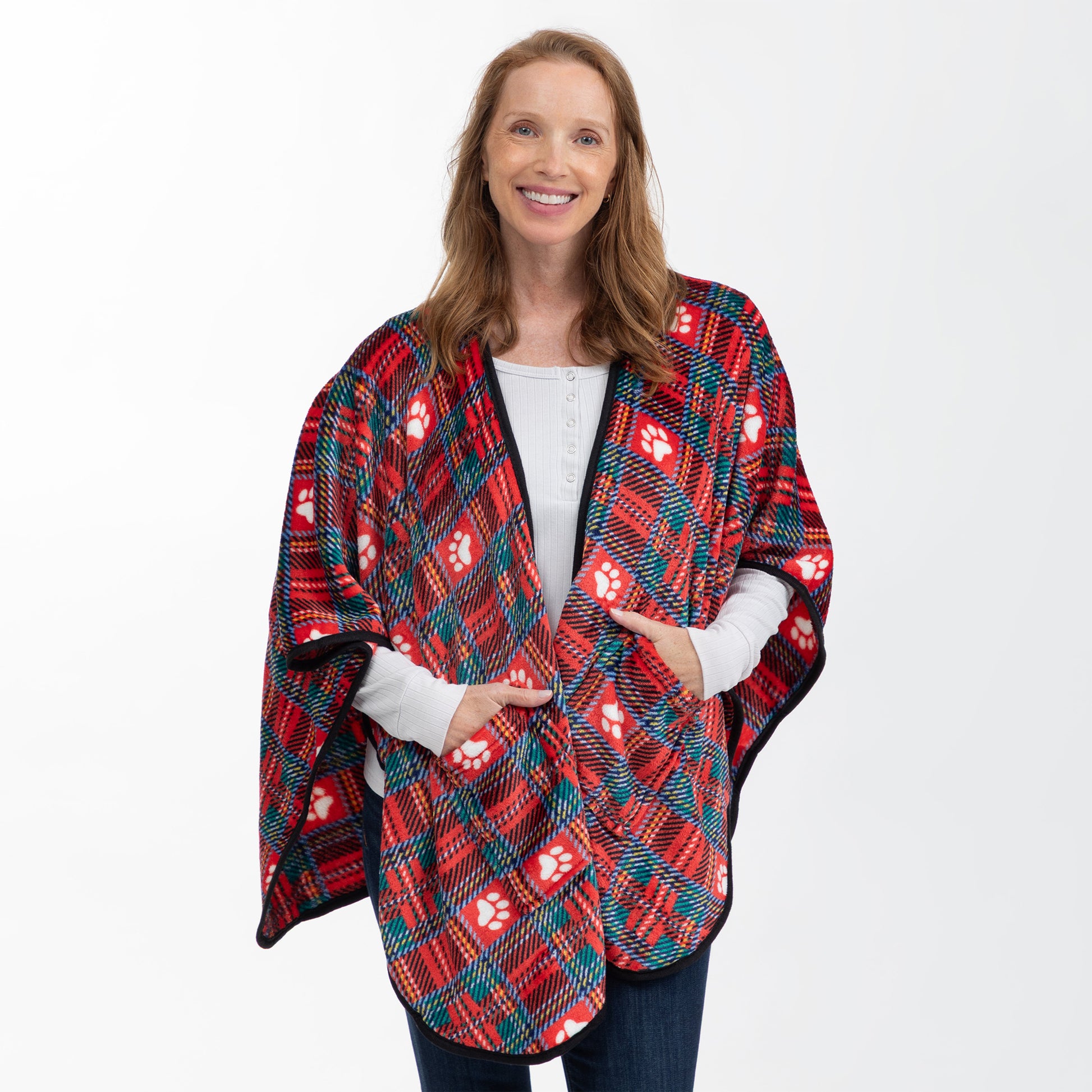 A smiling woman wearing a red, black, and teal plaid blanket poncho with white paw prints, styled over a white long-sleeve shirt and dark jeans, against a plain white studio background.
