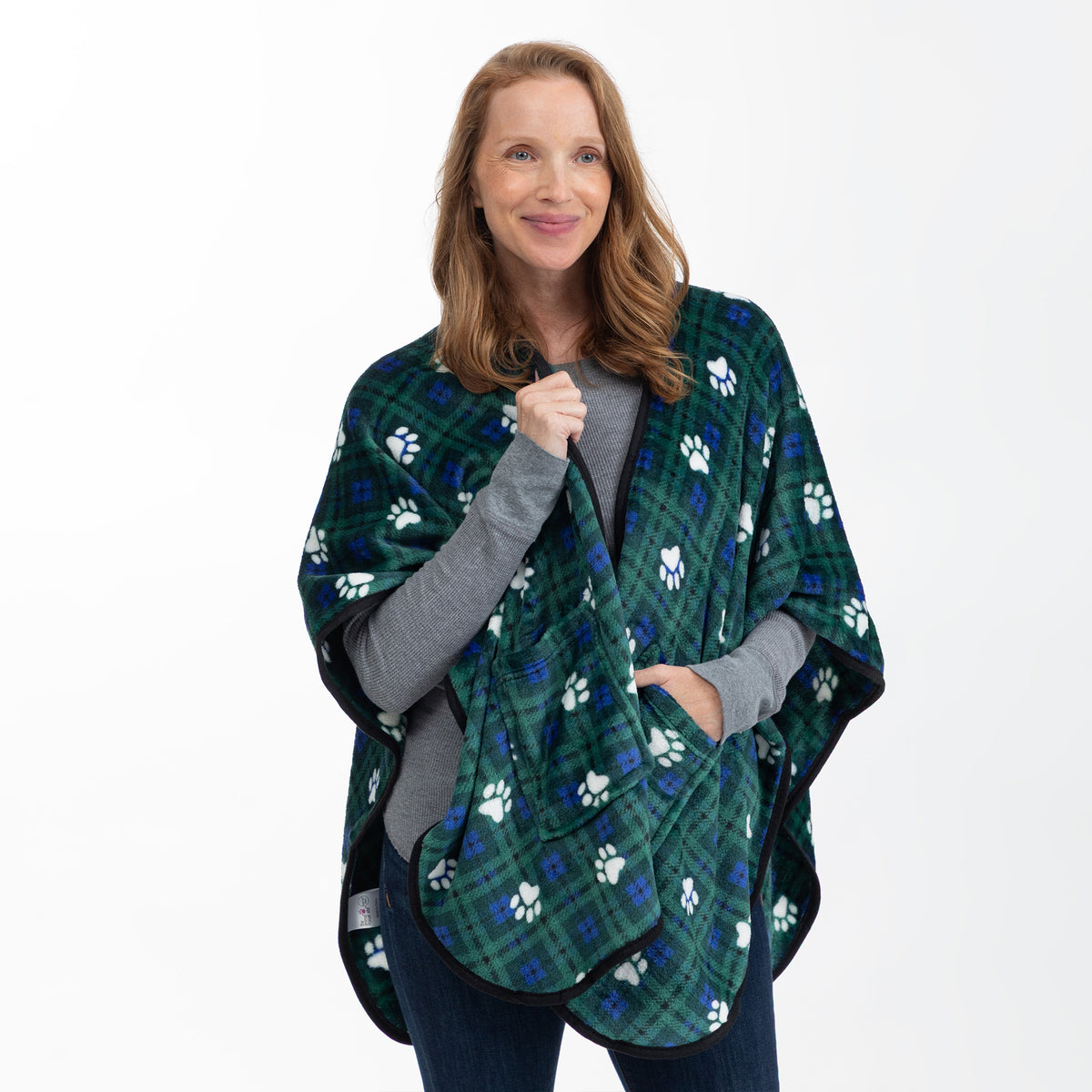 A woman modeling a green plaid shawl blanket with white paw prints, worn like a poncho over a gray long-sleeve shirt and blue jeans, against a plain white background.