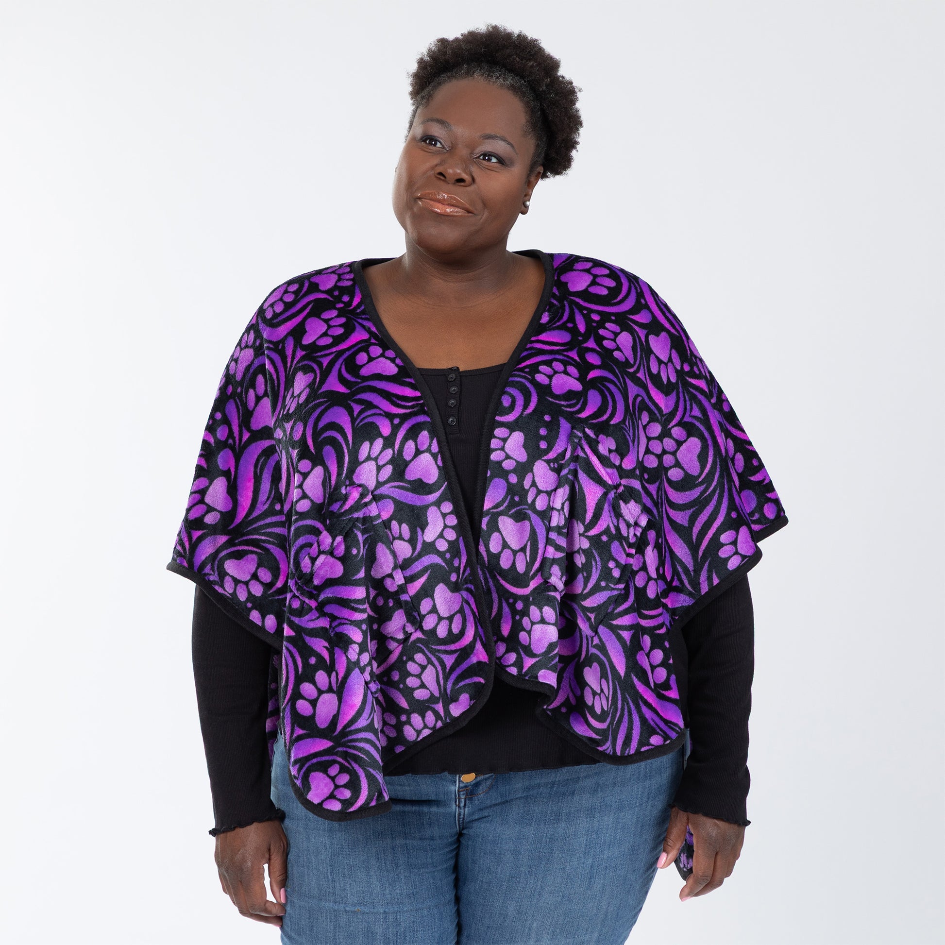 Woman wearing a fleece, pocketed shawl, patterned with purple paw prints and swirls and black piping, wrapped around her shoulders. Available in extended and plus size.