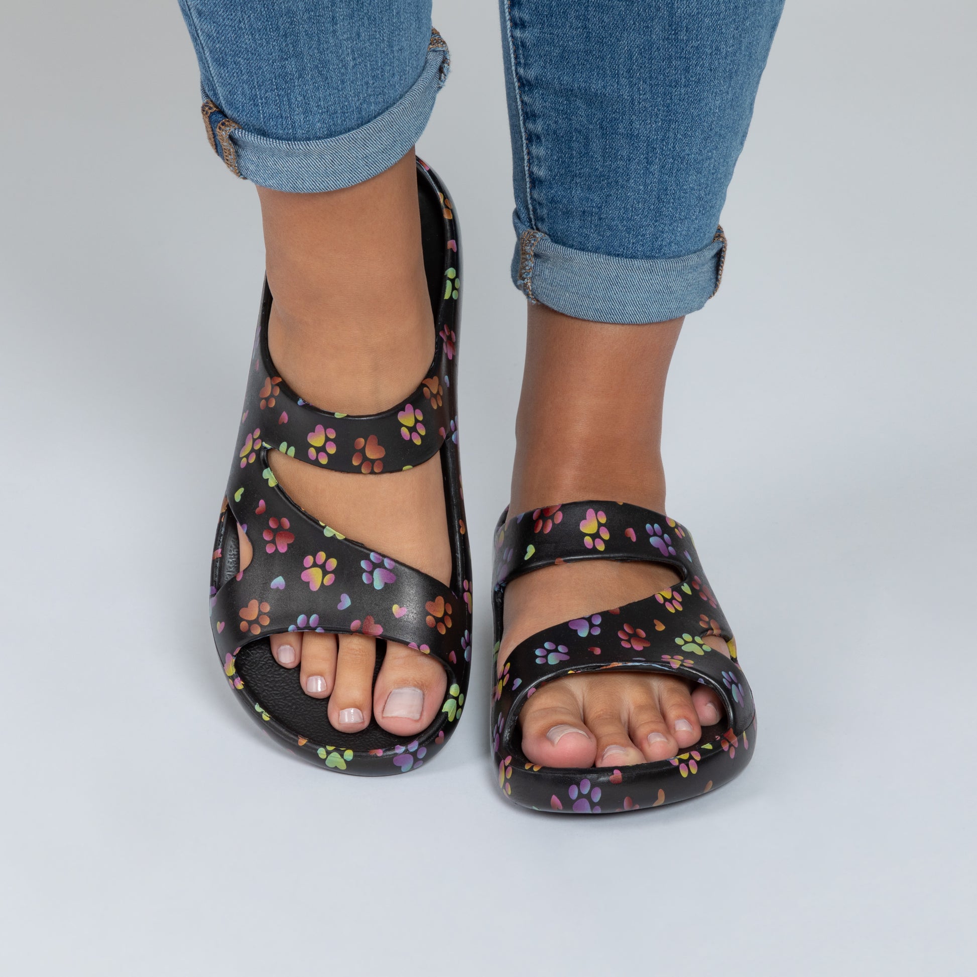 Black sandals with color paw print pattern worn with rolled-up jeans on a light gray background