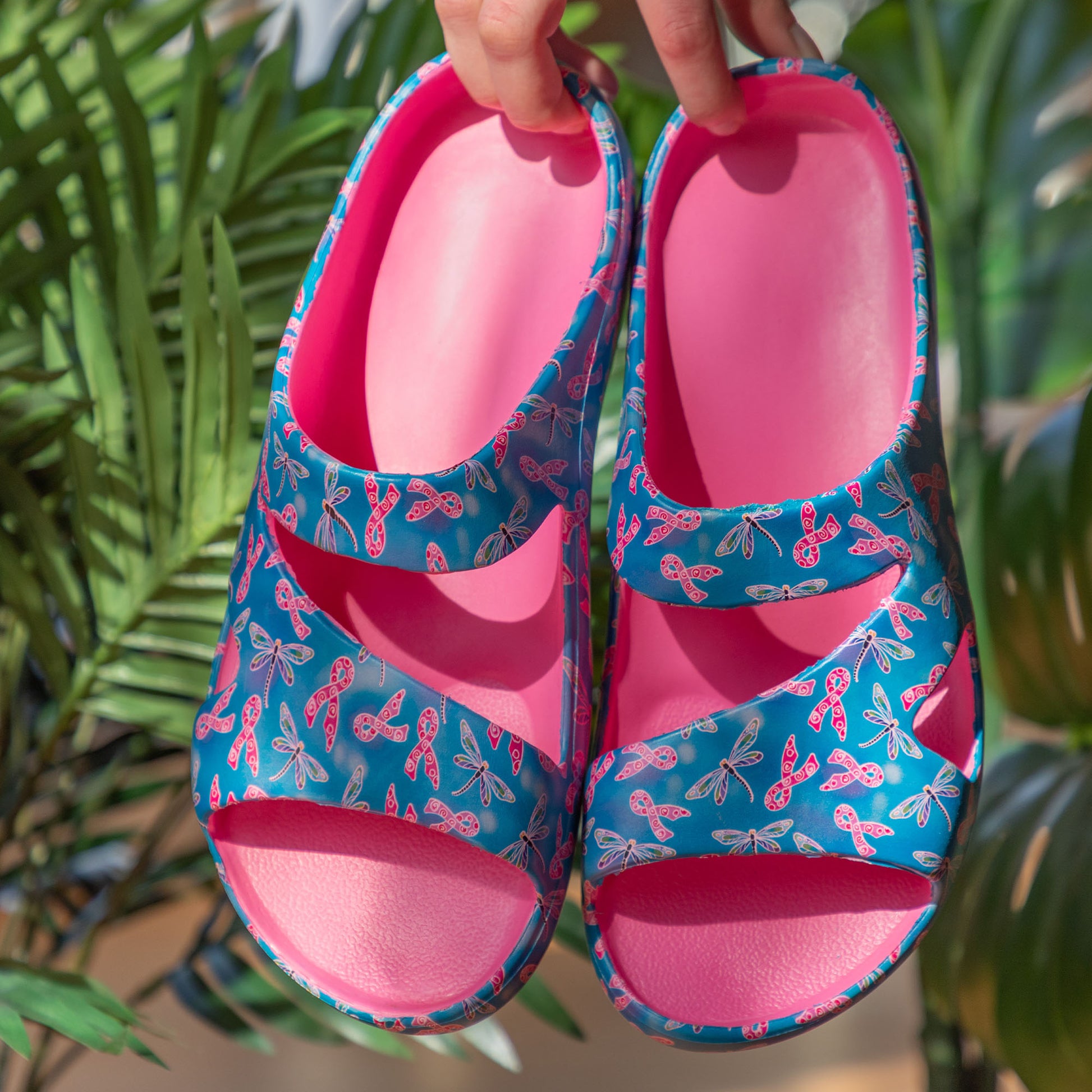 Pair of blue sandals with pink ribbon bows held against a green leafy background