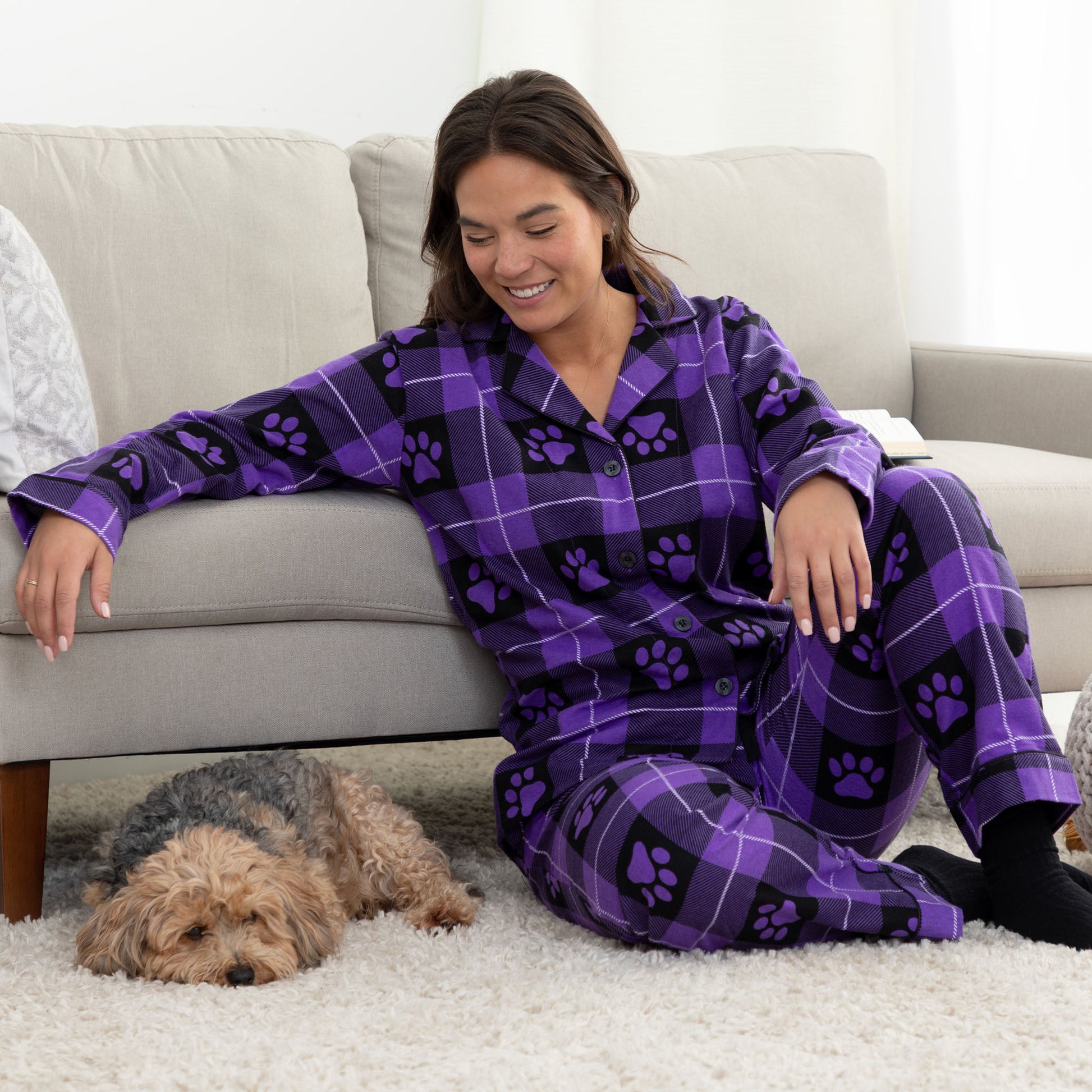 A woman sitting next to her pet dog in cozy living room setting. She is wearing a matching pajama set of purple, plaid, button-up, long-sleeve top patterned with paw prints and pants of the same color and pattern. 