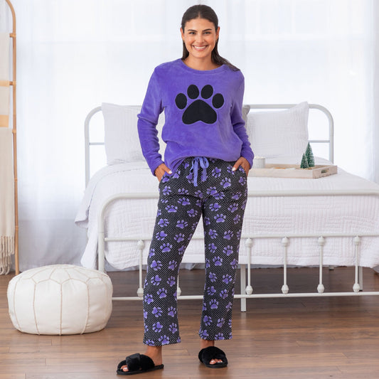 A woman standing in a bedroom wearing a purple long-sleeve shirt with a black paw print design on the center and a pair of pants designed with a black background and patterned with small white polka dots and purple paw prints.