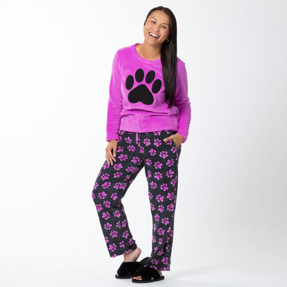A woman laughing while wearing a soft magenta long-sleeve shirt with a black paw print design on the center and a pair of soft black pants patterned with white polka dots and magenta paw prints. Her hand is in the pocket of the pants. 