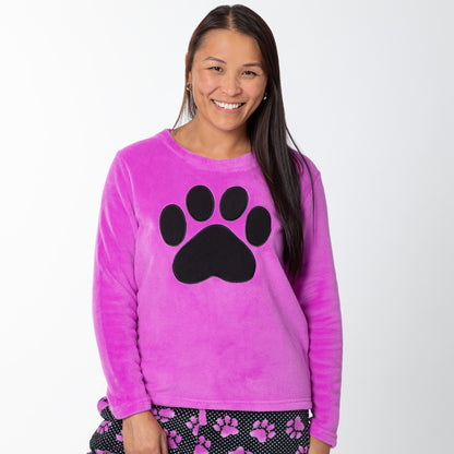 A woman wearing a soft magenta long-sleeve shirt with a black paw print design on the center and a pair of soft black pants patterned with white polka dots and magenta paw prints.