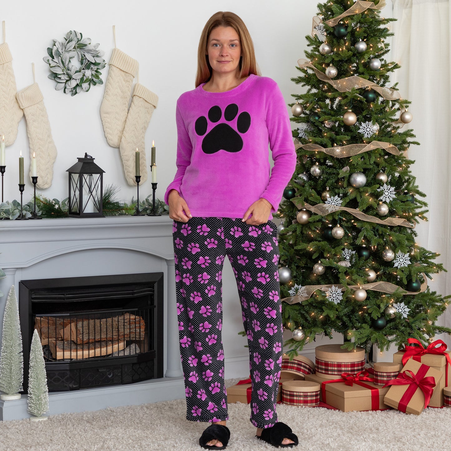 A woman standing in a living room setting with holiday decor. She is wearing a soft magenta long-sleeve shirt with a black paw print design on the center and a pair of soft black pants patterned with white polka dots and magenta paw prints.