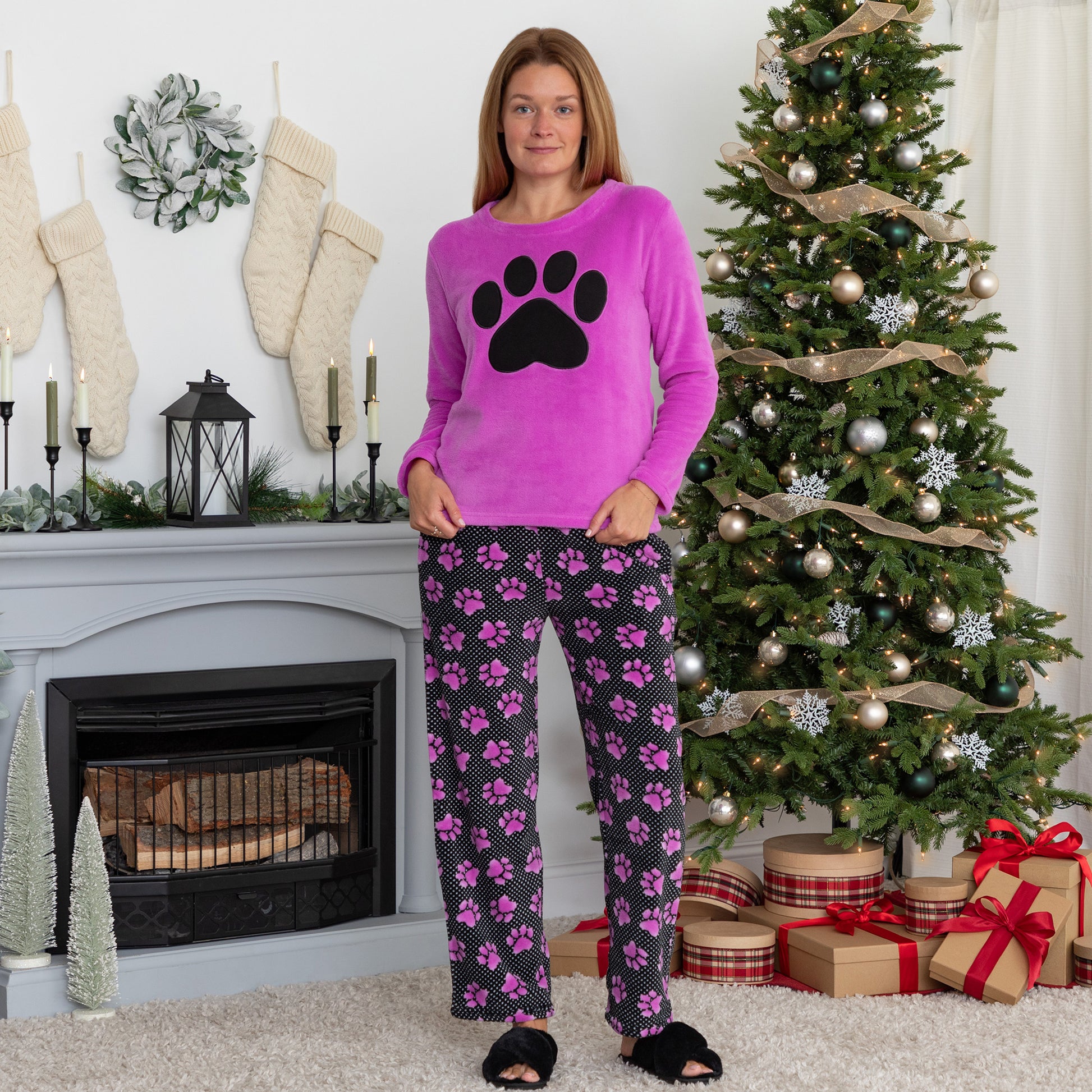 A woman standing in a living room setting with holiday decor. She is wearing a soft magenta long-sleeve shirt with a black paw print design on the center and a pair of soft black pants patterned with white polka dots and magenta paw prints.