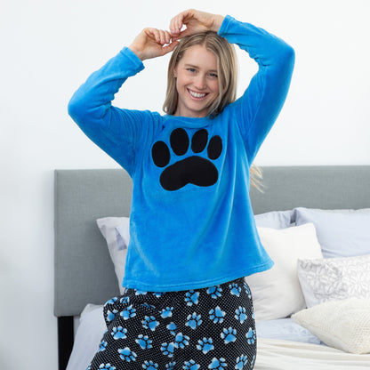 Woman wearing a blue fleece pajama top with a black paw print and matching pants in a bedroom.