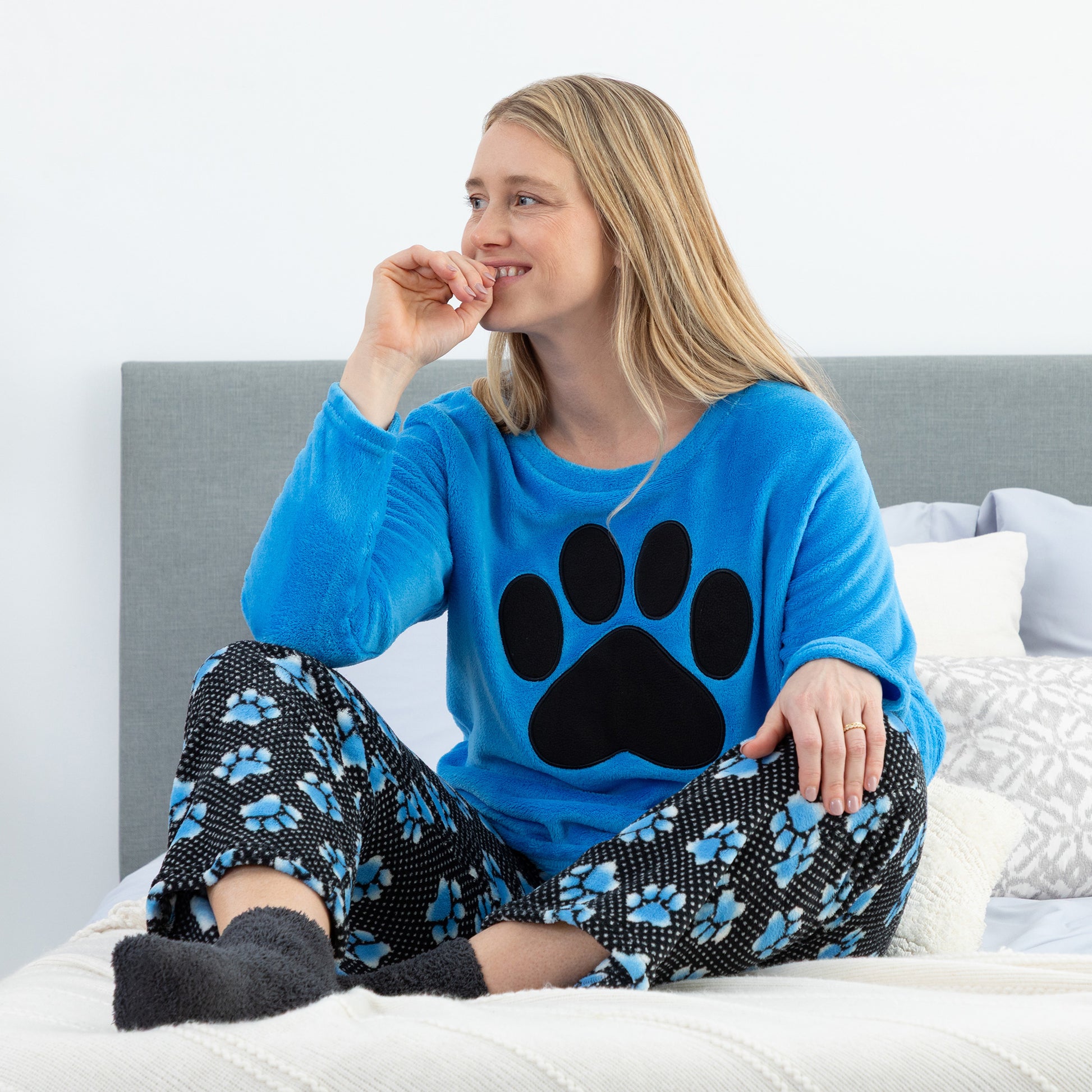Woman wearing a blue pajama top with a paw print and black pants with blue paw prints, sitting on a bed.