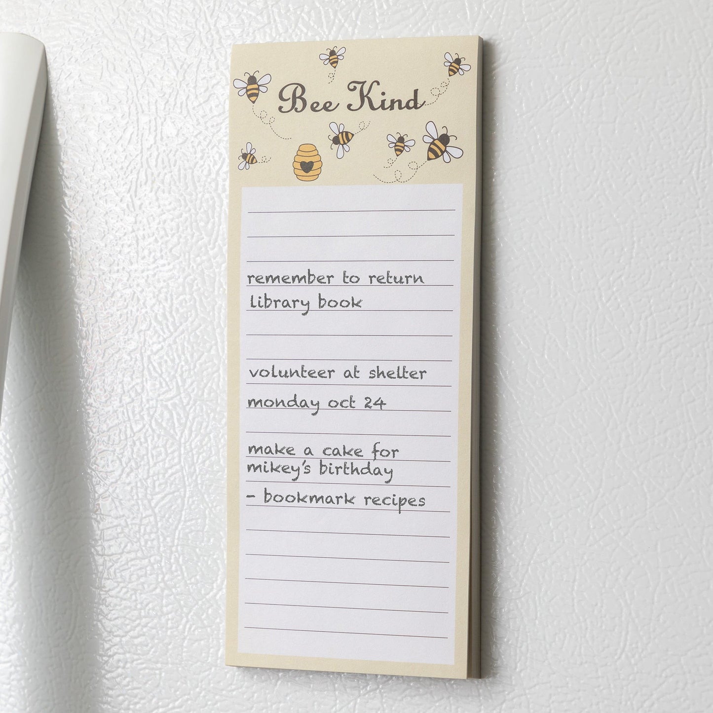 A light yellow notepad with bees and a honeycomb at the top and the phrase "Bee Kind" on a white refrigerator.