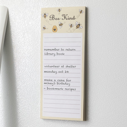 A light yellow notepad with bees and a honeycomb at the top and the phrase "Bee Kind" on a white refrigerator.