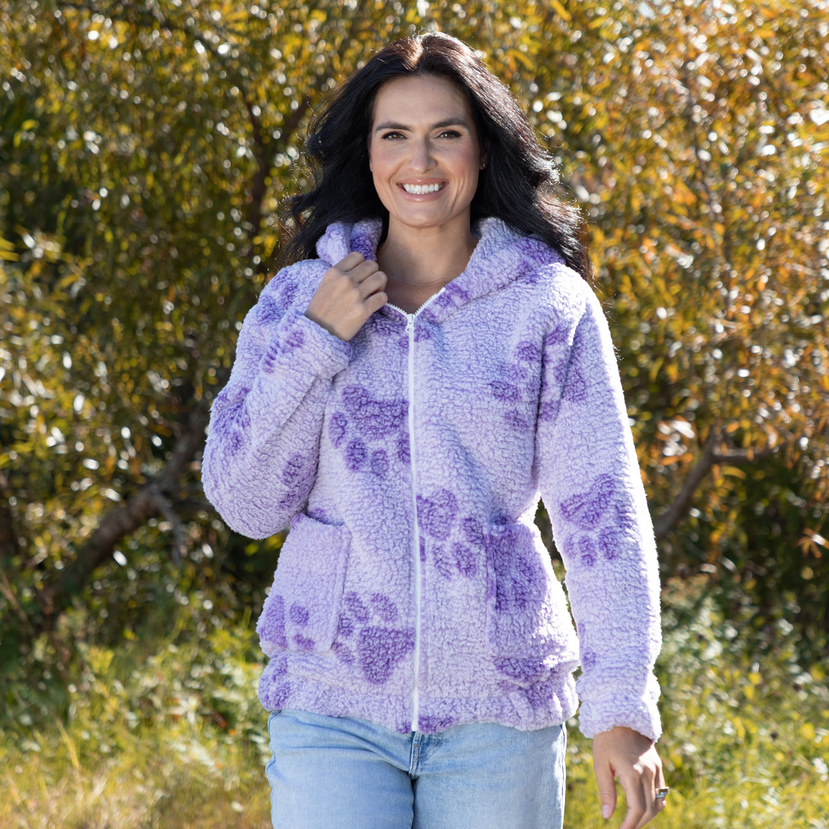 A woman wearing a light purple fuzzy zip-up hoodie with darker purple paw print patterns, standing outdoors in front of a blurred background of green and orange foliage.