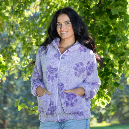 A woman wearing a light purple fuzzy zip-up hoodie with darker purple paw print patterns, standing outdoors in front of a leafy green tree.