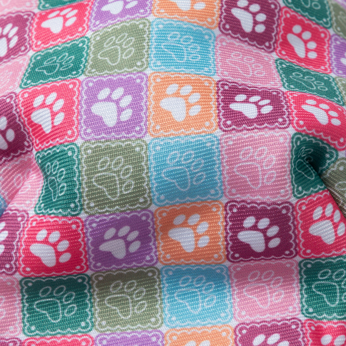 A close-up of a colorful fabric with a repeating pattern of square tiles, each containing a white paw print on a pink, purple, teal, green, and orange background.