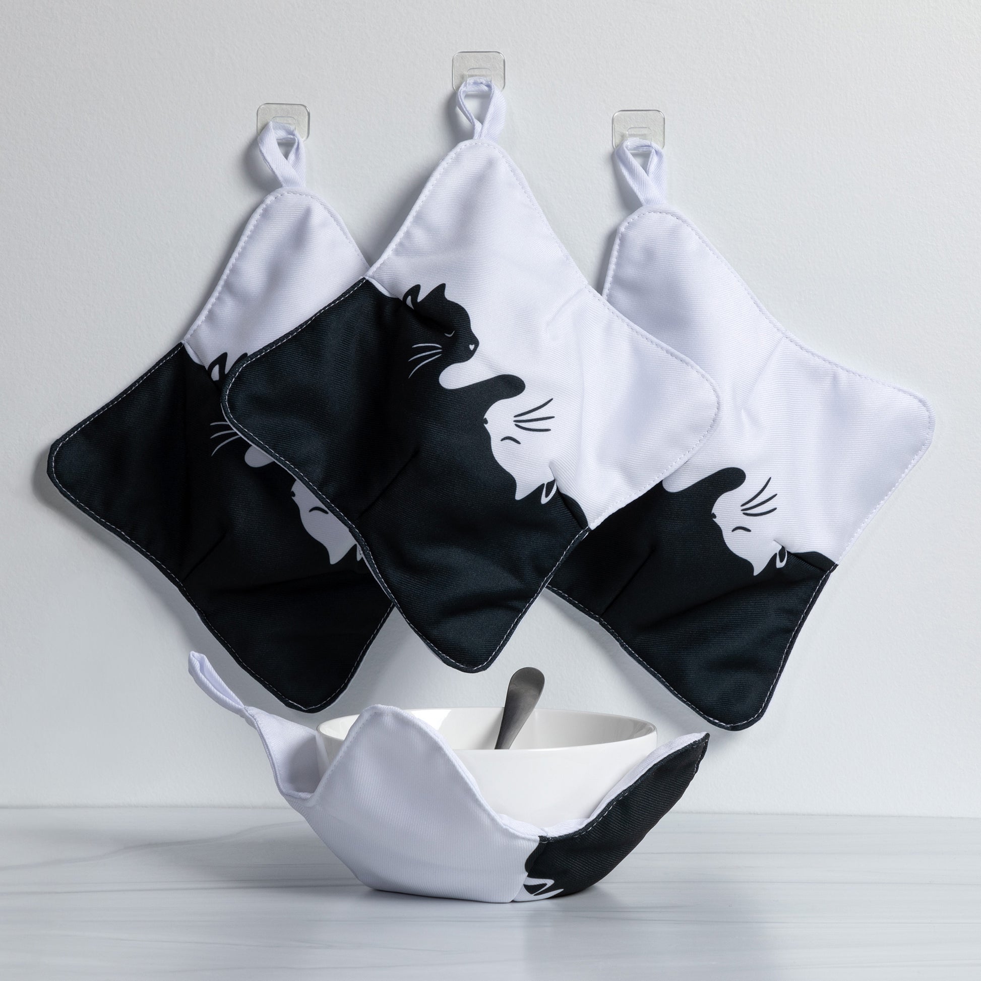 A set of four bowl cozies hanging from hangers in kitchen setting with one bowl sitting in a cozy, including a cat print design, snuggle cats