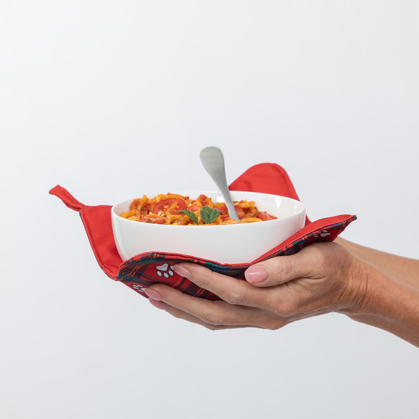 A white ceramic bowl filled with pasta and topped with a small green garnish, held in a red patterned potholder wrapped around two hands against a plain white background.