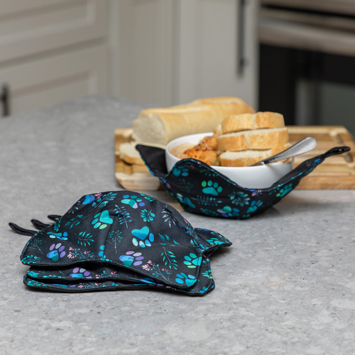 Two reusable fabric bowl cozies with a black background and blue, teal, and purple paw print pattern, shown on a kitchen counter with bread and a bowl of food in the background.