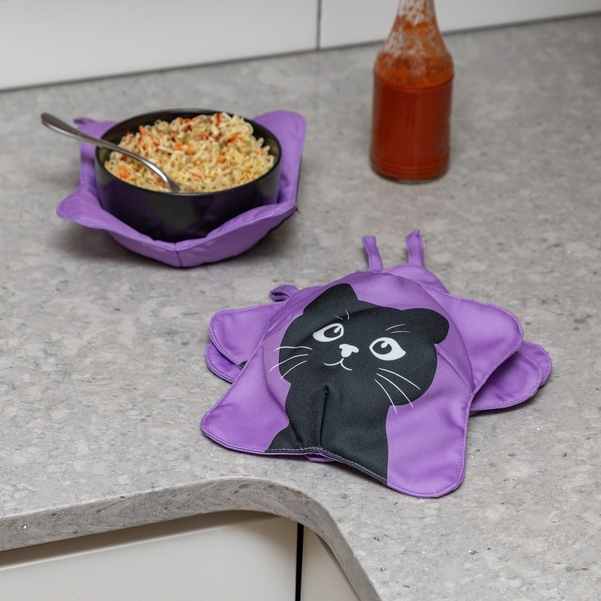 Purple bowl cozies on a gray kitchen countertop, one holding a black bowl of food and the other featuring a black cat face graphic.