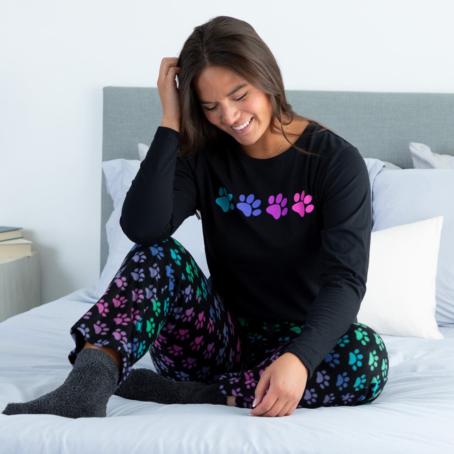 A woman sitting on a bed in a cozy bedroom wearing a black long-sleeve top with rainbow paw print and with design rainbow paws pants