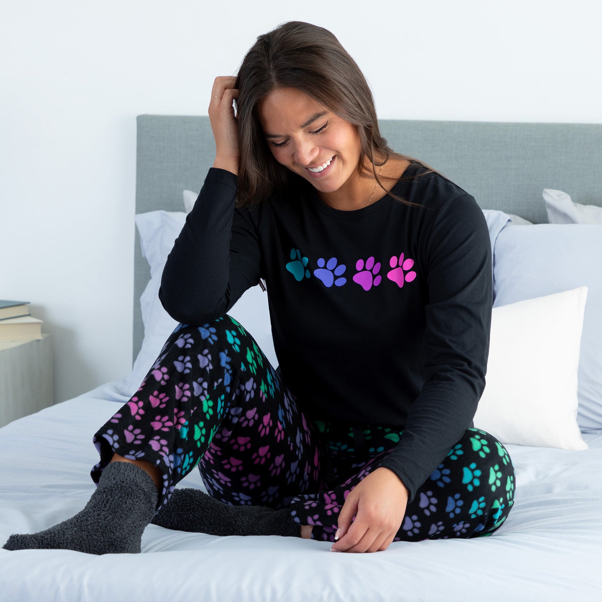 A woman sitting on a bed in a cozy bedroom wearing a black long-sleeve top with rainbow paw print and with design rainbow paws pants