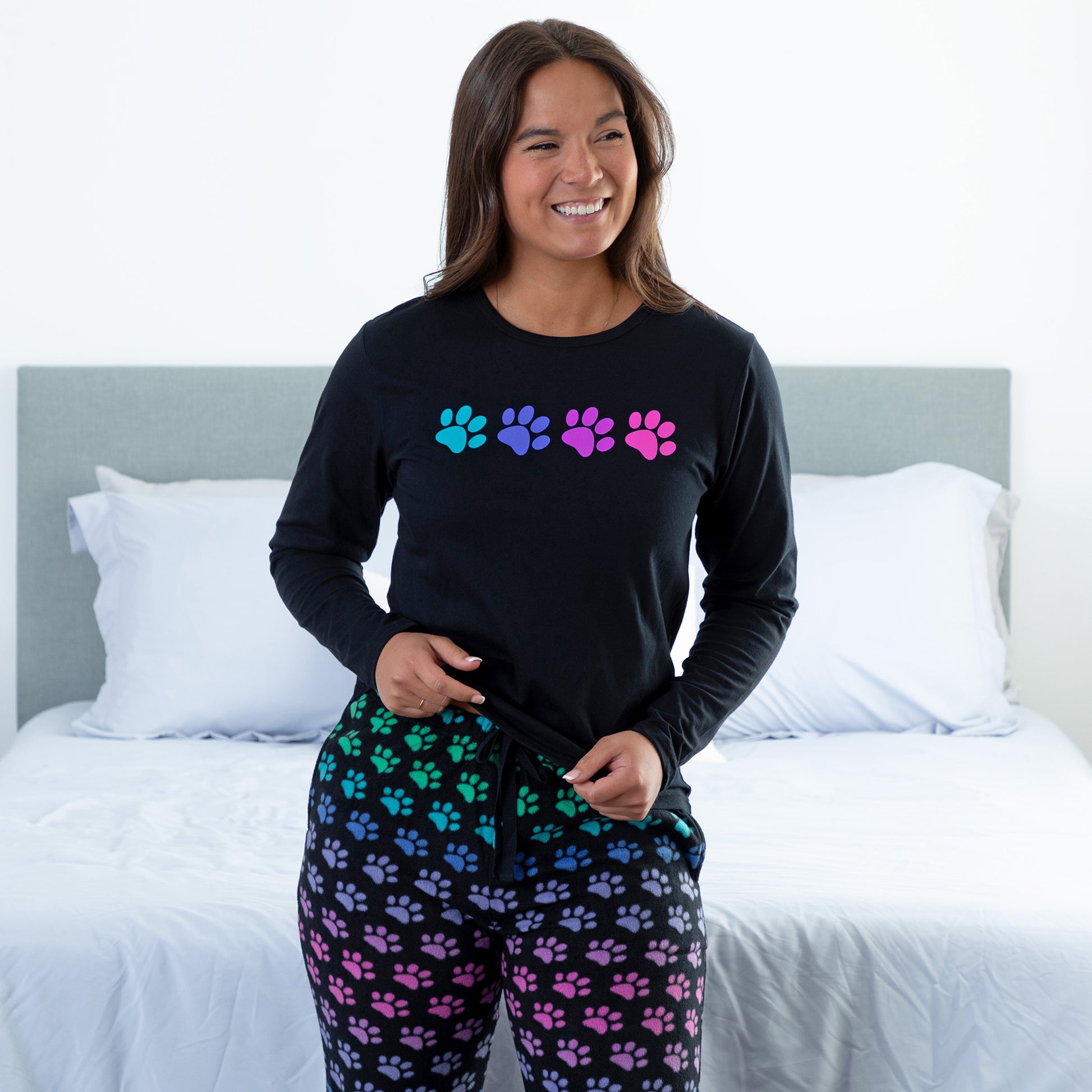 A woman standing in a bedroom wearing a black long-sleeve top with rainbow paw print and with design rainbow paws pants, slightly lifting the top show the drawstring adjustable waistband