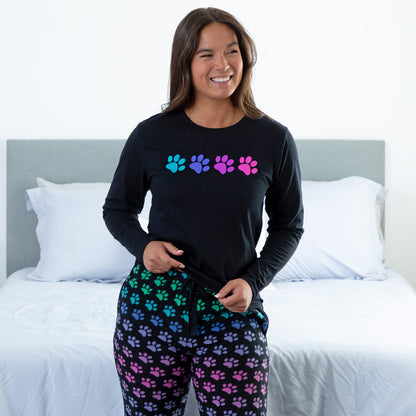 A woman standing in a bedroom wearing a black long-sleeve top with rainbow paw print and with design rainbow paws pants, slightly lifting the top show the drawstring adjustable waistband