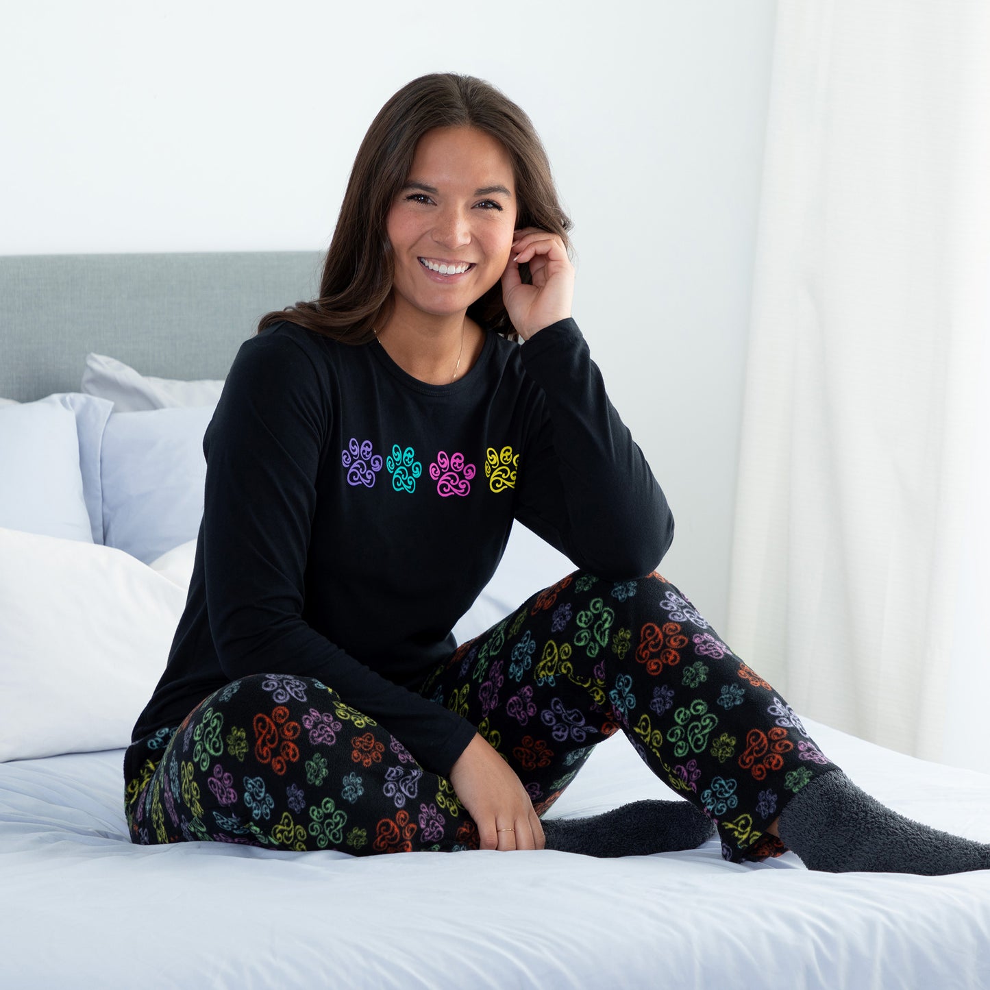 A woman sitting on a bed in a bedroom setting wearing a black long-sleeve top with swirling multicolored paw print and with design swirling multicolored paw print pants